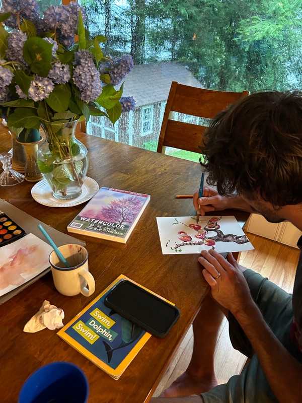 Tim painting at a table by a window, a jar of flowers and an open paint box beside him.
