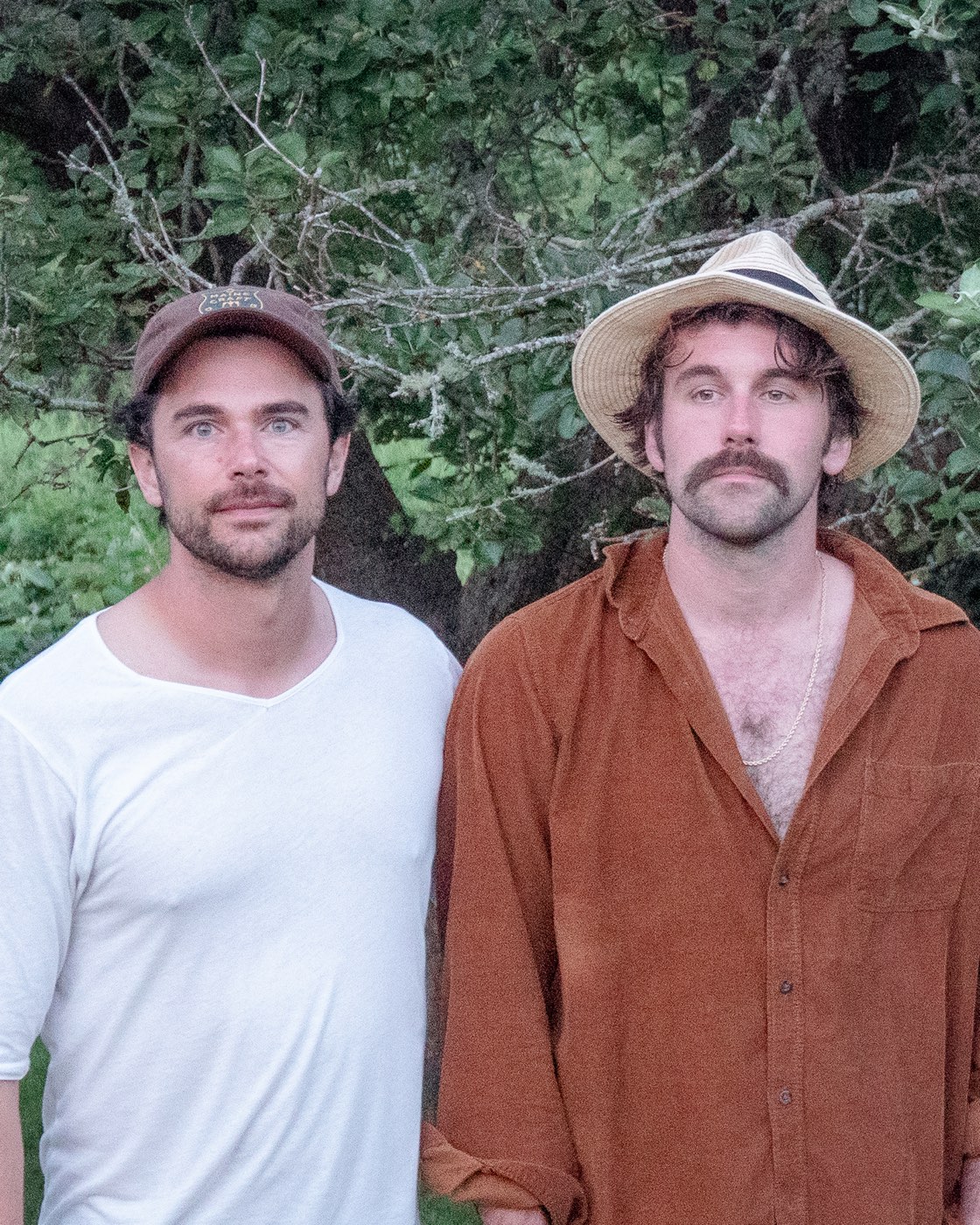 Tim Harrington and Paul Wright standing in front of dense green trees, one in a white shirt and cap, the other in a rust corduroy shirt and straw hat.